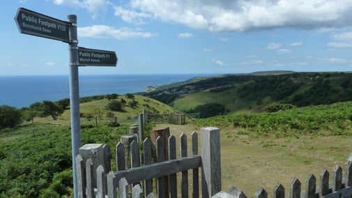 A signpost by a gate entices walkers to explore Ventnor Downs, with distant views to the sea way below in the distance